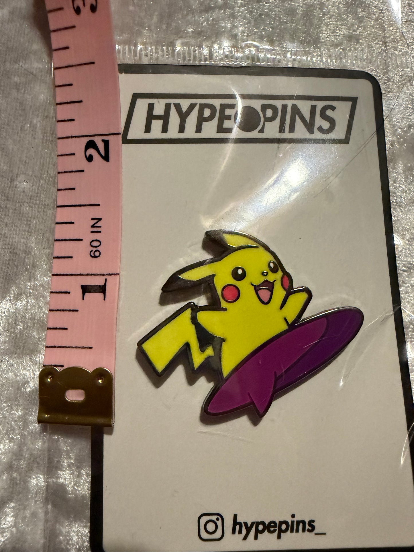 Surfing Pikachu Hard Enamel Pin by Hype Pins, displayed next to a tape measure to show its **1.5-inch size**. This dynamic Pokémon collectible features a durable hard enamel and black nickel metal finish.