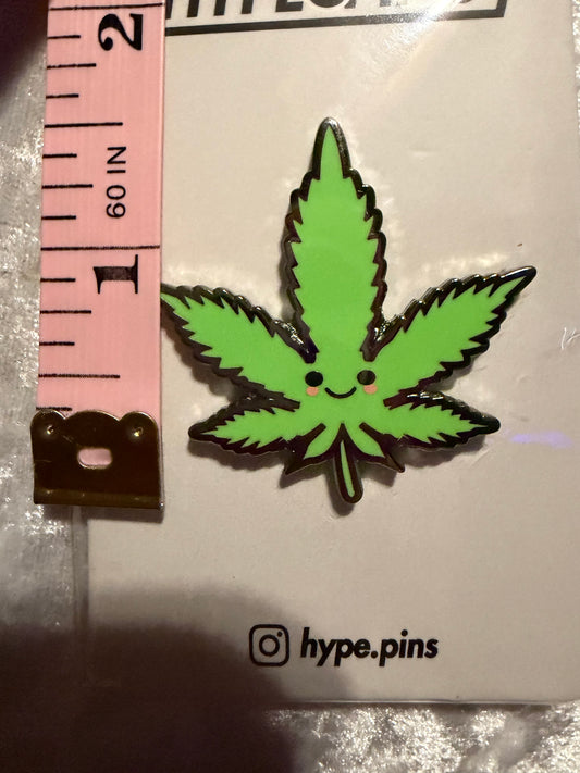 he 'Cute Weed Leaf' hard enamel pin displayed next to a common object (e.g., a quarter or a hand) to illustrate its compact, collectible size.