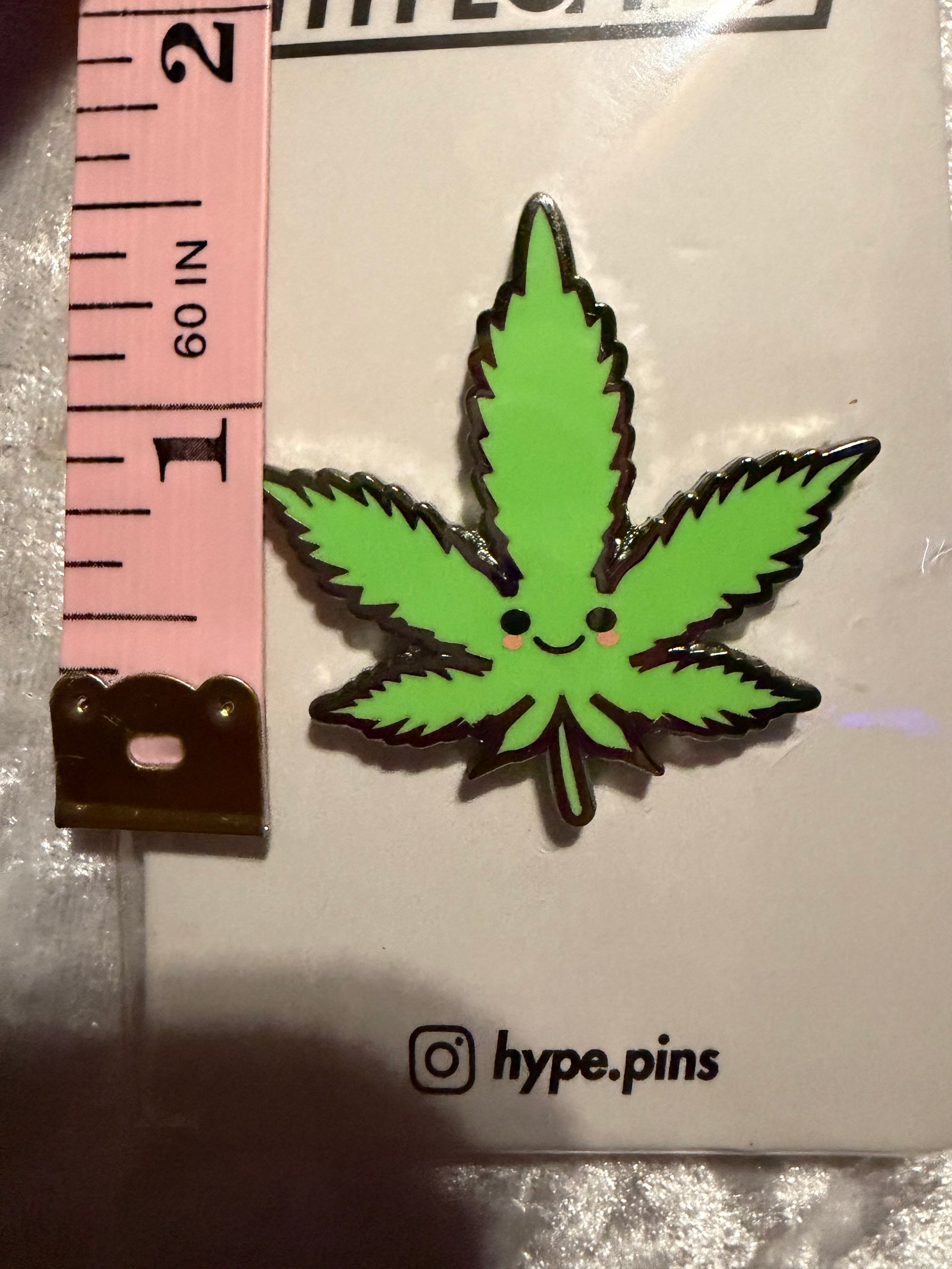 he 'Cute Weed Leaf' hard enamel pin displayed next to a common object (e.g., a quarter or a hand) to illustrate its compact, collectible size.